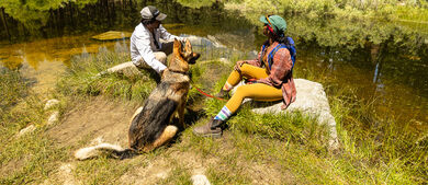 Two people and dog sitting next to river
